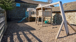 A wooden play structure with a slide and swings, set against a stone wall and a building with a sloped roof.