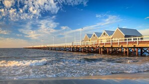 A pier extending into the sea with a row of buildings, a clear sky, and waves approaching the shore. - Apiwa Busselton Seaside Escape Apartments (Busselton)