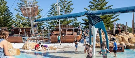 Un parque acuático con zona de juegos temática de barcos, piscina y árboles circundantes.