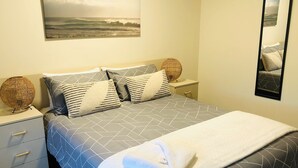 A neatly arranged bedroom with a bed, bedside tables, a mirror, and a wall-mounted artwork. - Apiwa Busselton Seaside Escape Apartments (Busselton)