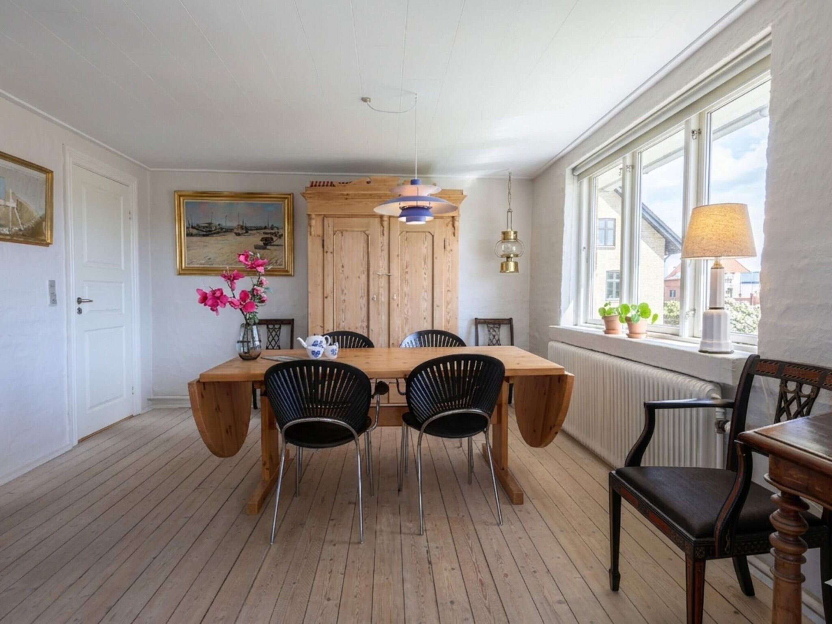 A dining area with a wooden table, four chairs, a vase with flowers, a painting on the wall, and a window with a view of buildings.