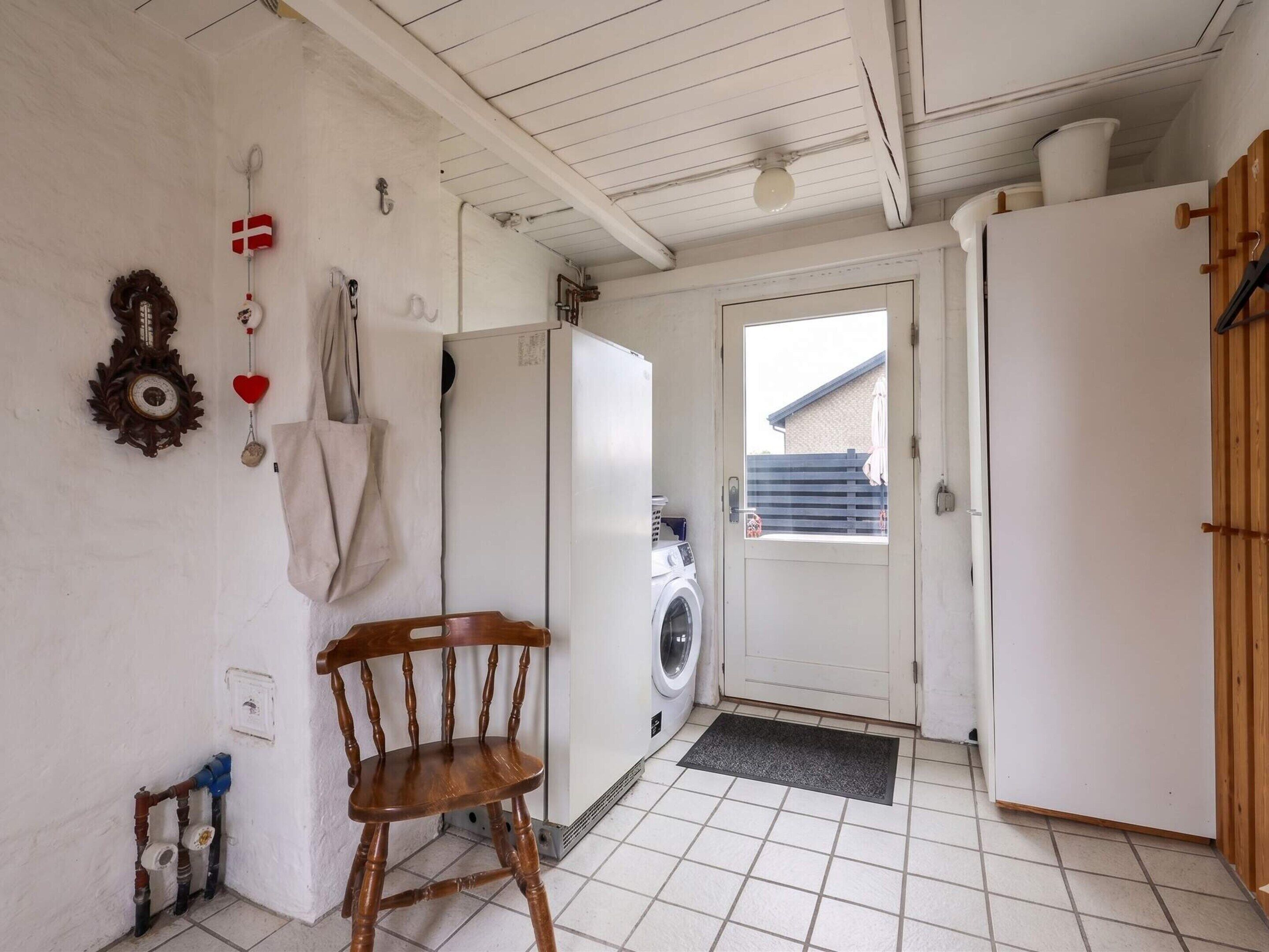 A laundry room with a washing machine, a wooden chair, a white door, and a small window.