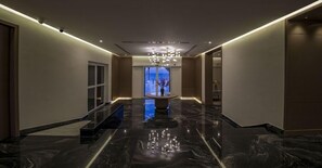 En modern hotellkorridor med ett blankt marmorgolv, en stor kristallkrona i mitten och utsikt genom ett stort fönster.