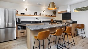 A modern kitchen with a stainless steel refrigerator, a built-in oven, a microwave, and a coffee machine. There is a wooden island with bar stools and pendant lighting. - Rock Creek Canyon | 1BR, Hot Tub, Sauna, Fire Pit (Twin Falls)