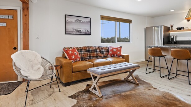 A modern living room with a sofa, a bench, and a chair. There is a kitchen area with a refrigerator and a coffee machine. A window provides a view of the outdoors. - Rock Creek Canyon | 1BR, Hot Tub, Sauna, Fire Pit (Twin Falls)