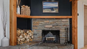 A stone fireplace with a wooden mantel, a framed landscape painting above it, and a stack of firewood to the left.
