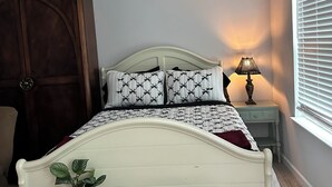 Un dormitorio con una cama, una mesita de noche, una lámpara, aire acondicionado, ventilador de techo y una ventana con persianas.