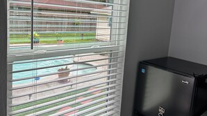 Habitación con ventana con persianas horizontales, un refrigerador negro y vistas a una piscina y edificios.