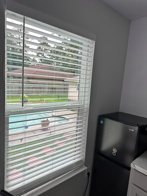 A room with a window with horizontal blinds, a black refrigerator, and a view of a swimming pool and buildings outside. - Private tiny home oasis with pool access. (Spring)