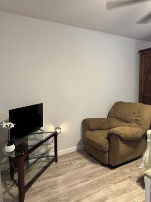 A living room with a flat-screen TV on a glass table, a brown armchair, and a white wall. - Private tiny home oasis with pool access. (Spring)