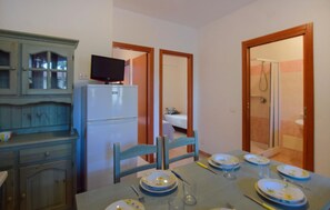Habitación compacta con una mesa de comedor lista para cuatro personas, un refrigerador pequeño, un televisor encima y un área de ducha visible a través de una puerta abierta.