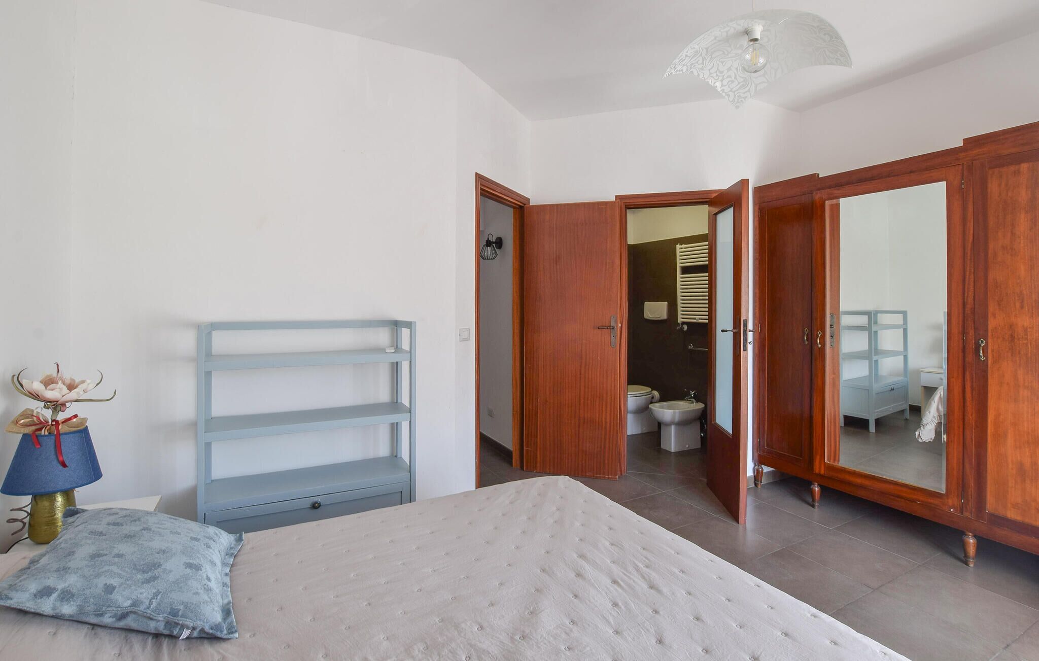 Een slaapkamer met een bed, nachtkastje, lamp, spiegel en een kledingkast.
