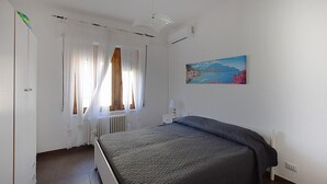 Een slaapkamer met een bed, een radiator, een raam met dunne gordijnen, een wandairconditioner en een schilderij.