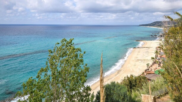 Une plage côtière aux eaux turquoise et limpides, avec un rivage sablonneux et une vue sur la mer qui s’étend jusqu’à l’horizon.
