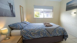 A bedroom with a bed, a nightstand, a window with blinds, and a wall-mounted light.