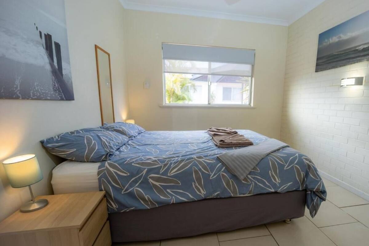 Un dormitorio con cama, mesita de noche, ventana con persianas y una lámpara fijada a la pared.