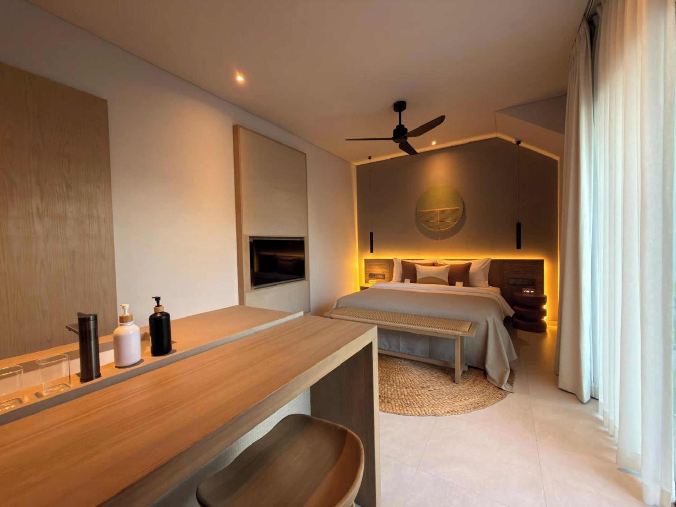 A modern hotel room with a wooden desk, a bed with a headboard, a ceiling fan, and a large window with curtains.
