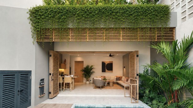 A modern outdoor pool area with a wooden deck, a seating area with a sofa and chairs, and a covered patio with plants. - Sin Nombre Homes  (Badung)
