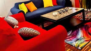 Una sala de estar con un sofá azul, almohadones coloridos, una mesa de centro, un ventilador de techo, un cuadro y una cortina llamativa.