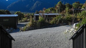 Uma entrada de automóveis de cascalho leva a um edifício com telhado de metal, cercado por vegetação e uma montanha ao fundo.