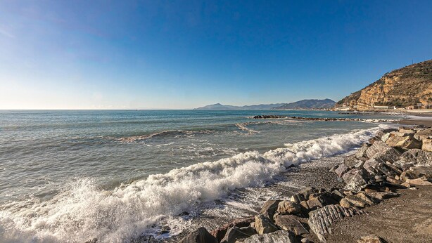Une côte rocheuse où les vagues s’écrasent sur le rivage, un ciel dégagé et des montagnes lointaines.