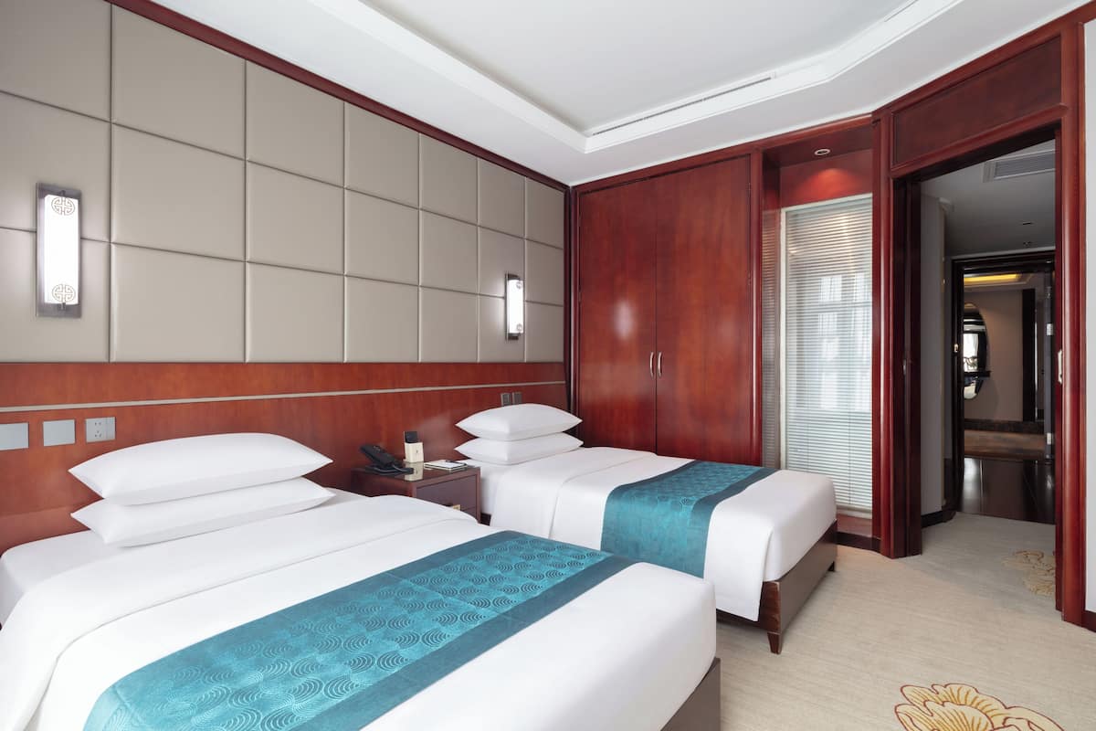 A hotel room with two beds, a wooden headboard, a bedside table, a nightstand, a mirror, and a door leading to a bathroom.