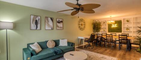 A living room with a green sofa, a coffee table, a dining area with chairs, a ceiling fan, and wall art.