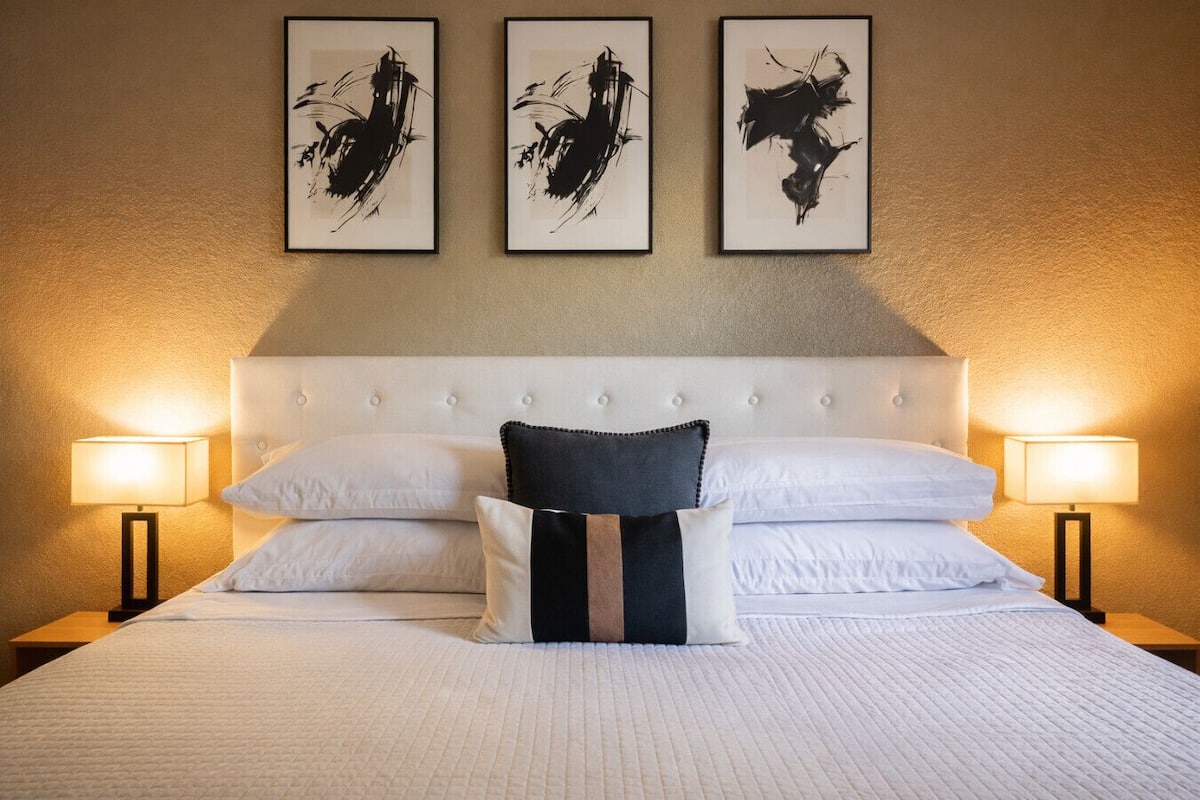 Una habitación de hotel con una cama grande, ropa de cama blanca, un cabecero capitonado y tres cuadros abstractos blanco y negro en la pared.