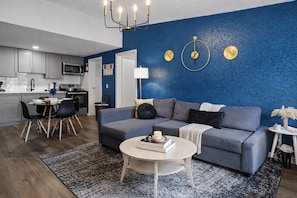 Una sala moderna con una pared azul de acento, un sofá gris, una mesa de centro redonda y una zona de comedor con una mesa y sillas blancas.