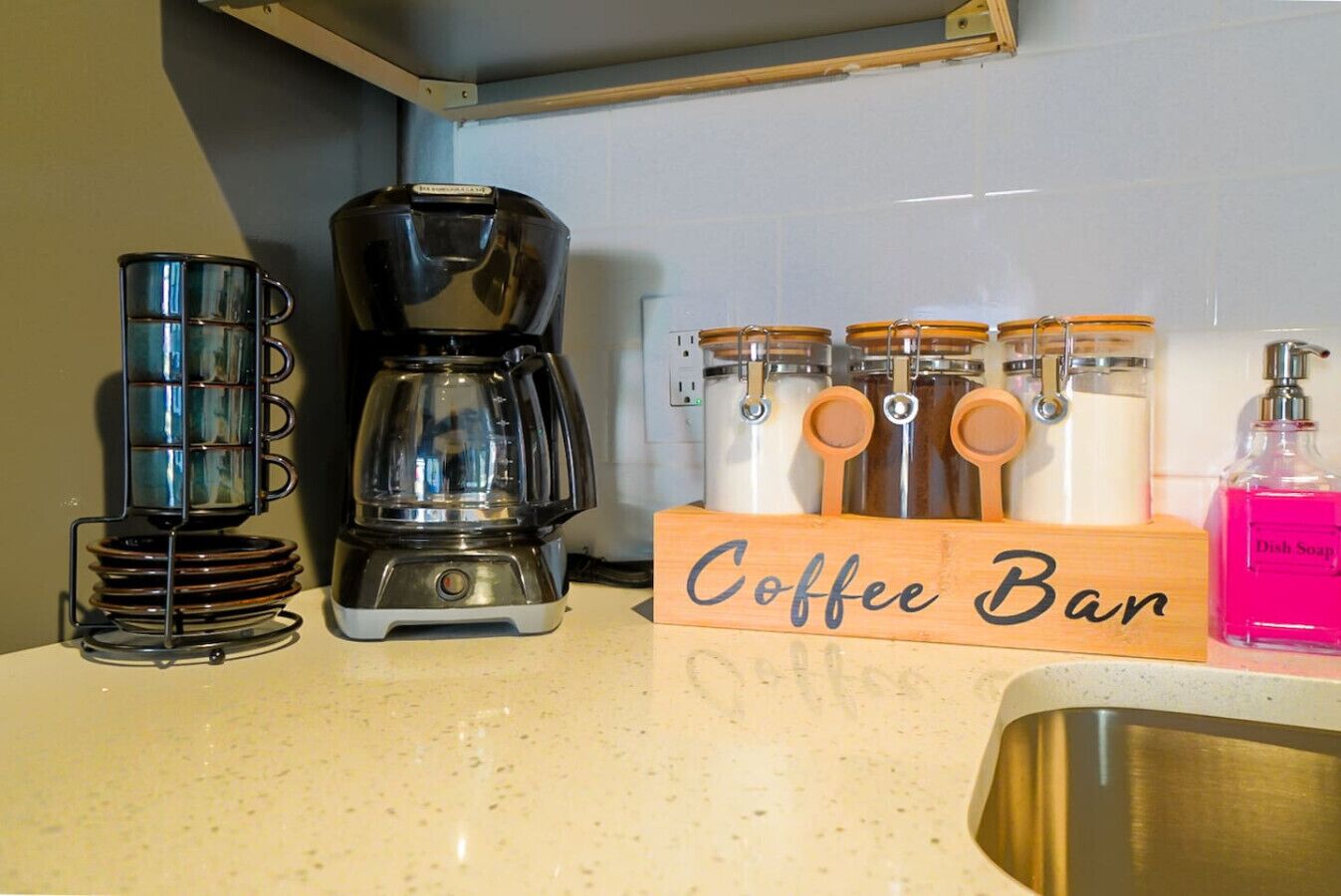 Una encimera de cocina con una cafetera, una taza de café, un letrero de cafetería, frascos y una botella de detergente.