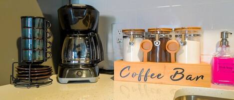 Una encimera de cocina con una cafetera, una taza de café, un letrero de cafetería, frascos y una botella de detergente.