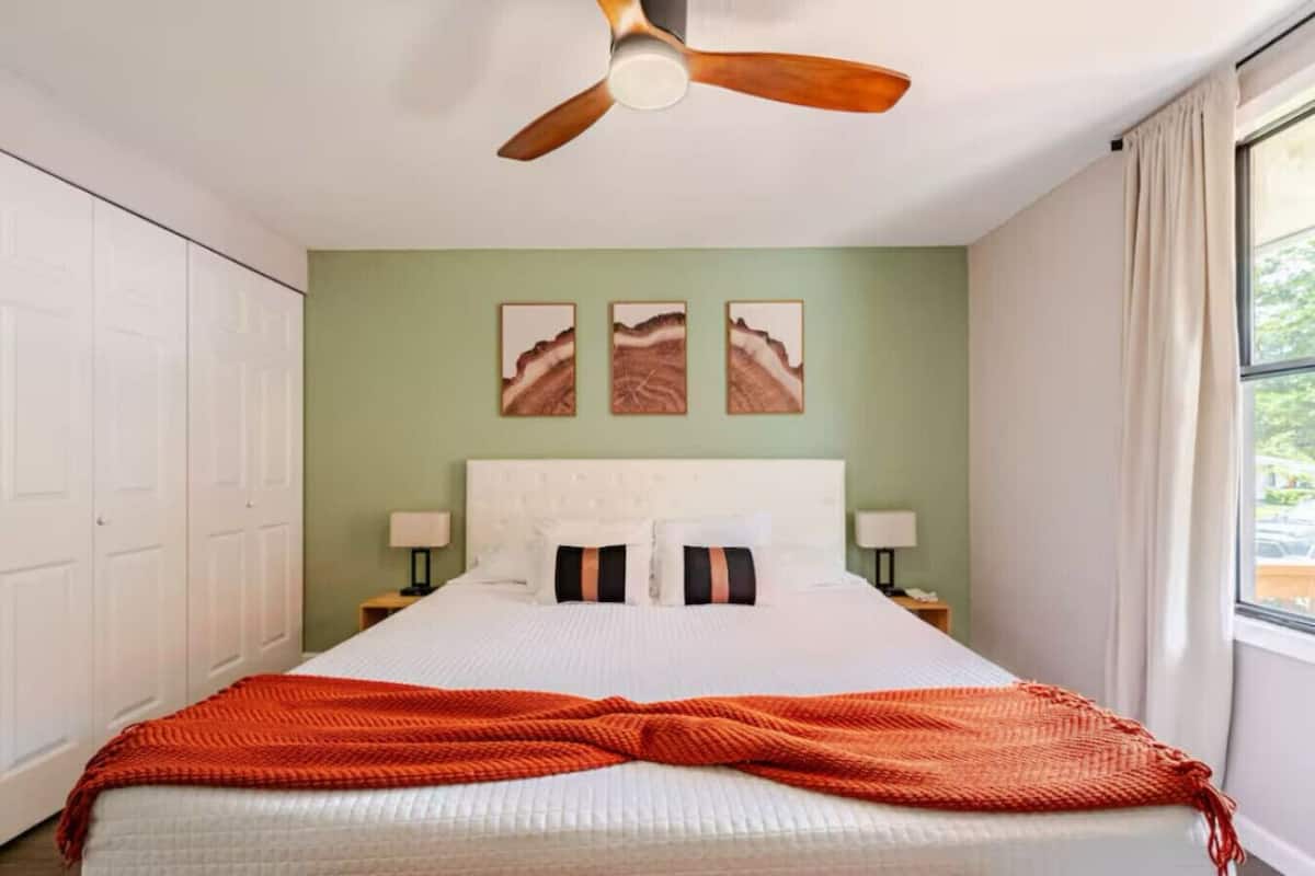 Un dormitorio con cama, ventilador de techo, ventana y cuadros en la pared.