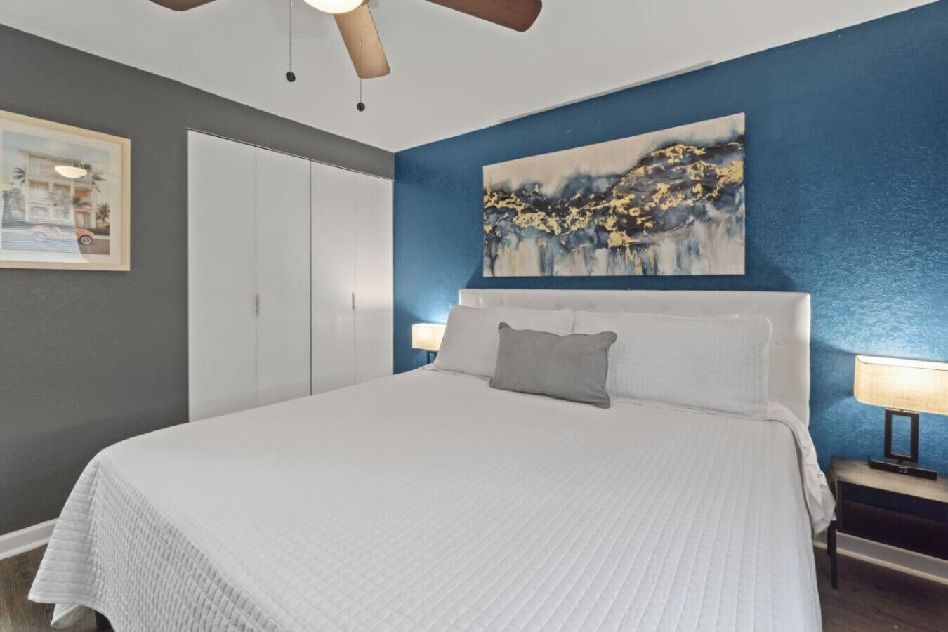 A bedroom with a large bed, a ceiling fan, a painting, and a wardrobe.
