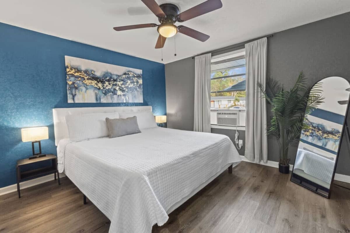 Un dormitorio con una cama grande, ventilador de techo, un cuadro en la pared, ventana con cortinas y una planta.