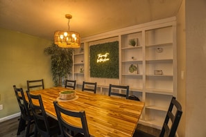 A dining area with a wooden table, chairs, a chandelier, a plant, and a neon sign.