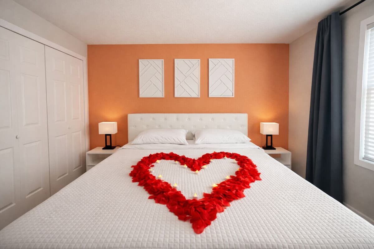 Una habitación de hotel con una cama grande, ropa de cama blanca, una decoración con forma de corazón hecha de flores rojas, dos lámparas de noche y una ventana con cortinas.