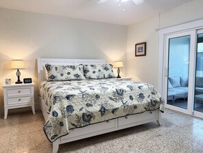 Ein Schlafzimmer mit einem Bett, zwei Nachttischen, einer digitalen Uhr, einem gerahmten Bild und einer Glasschiebetür, die auf einen Balkon führt.