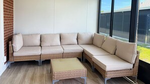 Een zithoek met een rieten bank en een bijpassend poef, een groot raam met uitzicht op een gebouw, en een bakstenen muur.