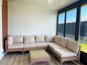Eine Sitzecke mit Winkelsitz, einem Korbsessel und passendem Hocker, ein großes Fenster mit Blick auf ein Gebäude und eine Ziegelwand.