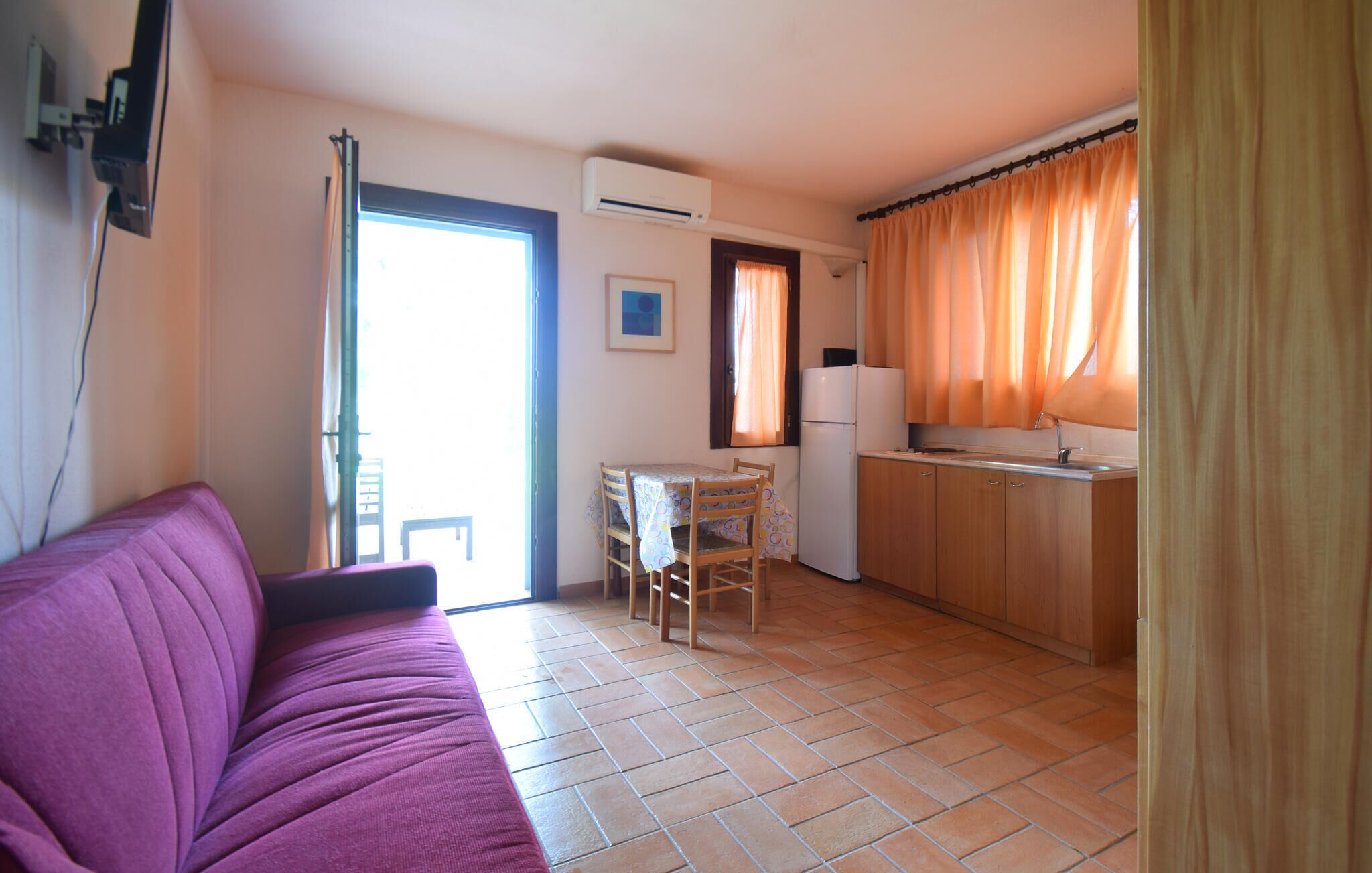 A compact living space with a purple sofa, a dining table with chairs, a kitchen area with a refrigerator, and a sliding glass door leading to an outdoor area.