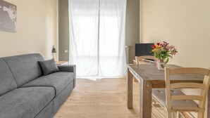 Una habitación con un sofá gris, una mesa de comedor de madera con dos sillas, una cortina blanca y una pequeña mesa con un florero.