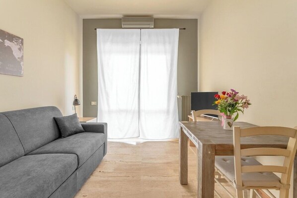 A room with a grey sofa, a wooden dining table with two chairs, a white curtain, and a small table with a vase of flowers. - Residence Fraschini (Milan)