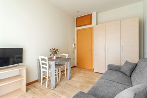 Una sala de estar moderna con un sofá gris, un televisor sobre un mueble, una mesa de comedor con sillas y un armario.