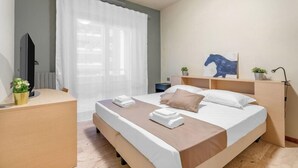 Habitación de hotel con cama, escritorio, radiador, ventana con cortinas, televisor montado en la pared y una planta pequeña.
