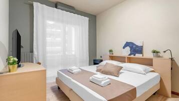 A hotel room with a bed, a desk, a radiator, a window with curtains, a wall-mounted TV, and a small plant.