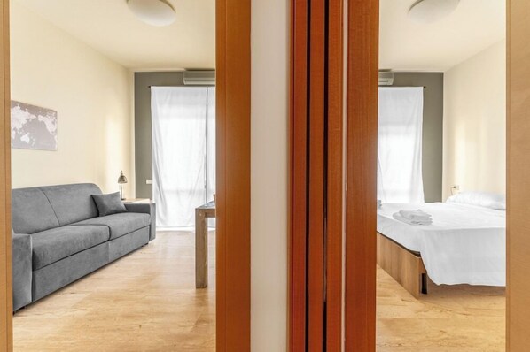 A modern hotel room with a grey sofa, a wooden bed frame, and a small table. - Residence Fraschini (Milan)