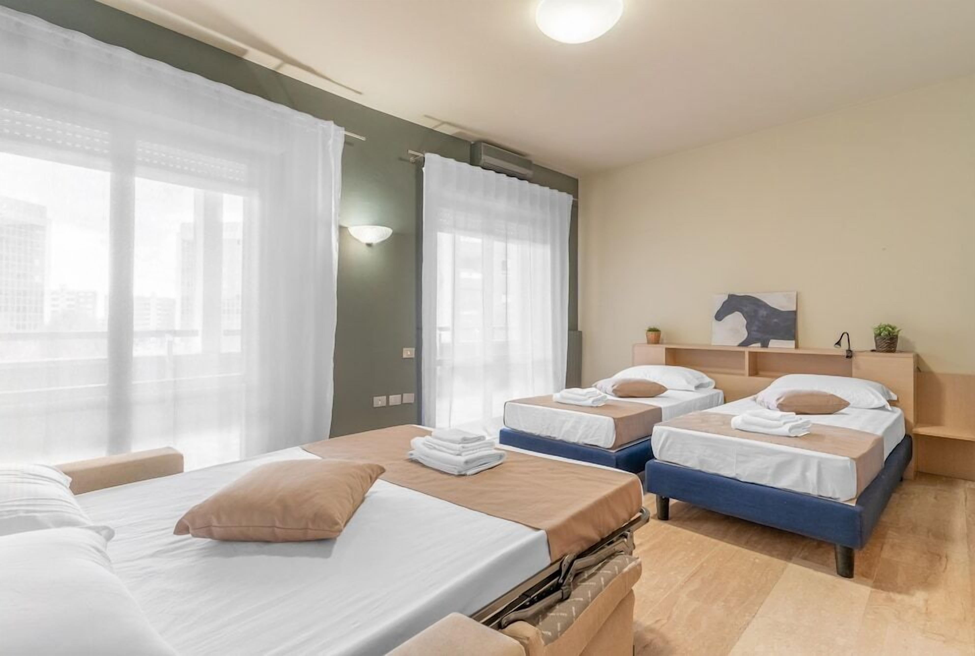 A hotel room with two beds, a large window with sheer curtains, a small desk, and a wall-mounted shelf with a plant.