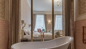 Una habitación con una bañera independiente grande, una cama con cubrecama blanco y un espejo montado en la pared que refleja la habitación.