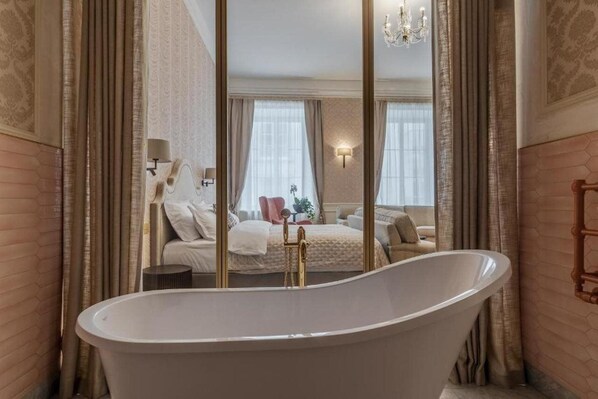 Ein Zimmer mit einer großen, freistehenden Badewanne, einem Bett mit weißer Tagesdecke und einem an der Wand befestigten Spiegel, der den Raum reflektiert.