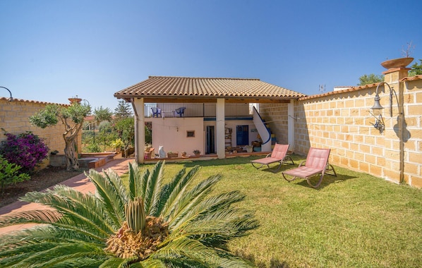 Une maison à deux étages, avec un toit en tuiles, une terrasse couverte avec des chaises et un petit jardin agrémenté de palmiers.